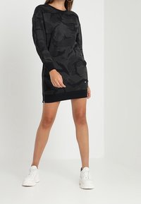 Musta kamouflage-kuosisweatshirt-mekko, jossa on pitkät hihat, ribbireunat hihoissa ja helmassa, sivuvetoketjuyksityiskohtia ja rento leikkaus.