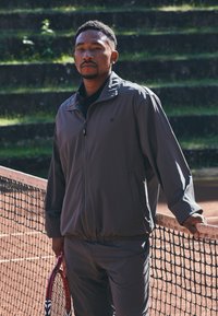 Mann i grå treningstøy som holder en rød tenniskølle, står ved nettet på en utendørs leir tennisbane med gress terrasser i bakgrunnen.