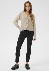 Beige knitted cardigan with a deep V-neck, featuring three large brown buttons and blue accents on the cuffs, paired with black skinny jeans.