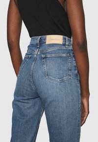 Modré džíny s vysokým pasem, které mají hnědou koženou etiketu s nápisem "Calvin Klein", dvě zadní kapsy a rovný střih.