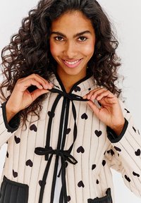 Béžová prešívaná bunda s vertikálnymi čiernymi pruhmi a rozptýlenými čiernymi srdcovými vzormi. Má kapucňu na šnúrku a dva predné vrecká.