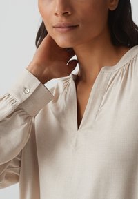 Beige blouse gemaakt van lichtgewicht stof, met een V-hals, pofmouwen en een knoopdetail op de manchet met een subtiele textuur.