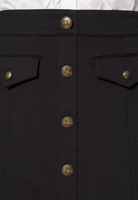 Svart kjol med framknappar och två övre fickor. Guldfärgade detaljer med en texturerad yta. Materialet verkar robust och välsytt.