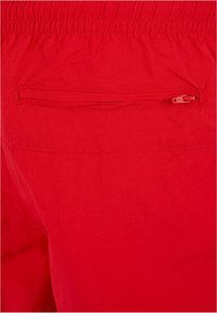 Pantaloni scurți roșii dintr-un material ușor, cu o talie elastică și un buzunar la spate cu fermoar. Textură fină fără modele vizibile.
