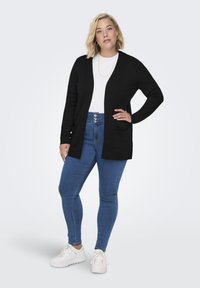 Zwarte open voorkardigan met lange mouwen, twee voorkruimelnaden, gedragen over een witte top en gecombineerd met blauwe skinny jeans en witte sneakers.