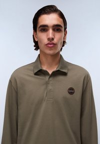 Olijfgroene polo met lange mouwen en een knoopsluiting bij de kraag, voorzien van een ronde logopatch op de linkerborst en een gladde stoftextuur.