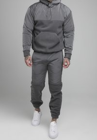 Felpa grigia con cappuccio e tasca frontale, abbinata a pantaloni jogger grigi con pannelli testurizzati. Sneakers bianche completano l'outfit.