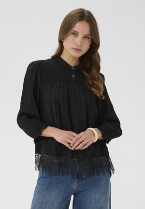 Woman with long brown hair wears a black blouse with lace hem and blue jeans, posing with hands clasped and a white bracelet on wrist.