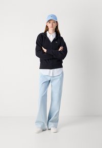 Sudadera negra con cremallera, camisa blanca con cuello, jeans baggy azul claro y zapatillas blancas. Gorra azul claro con logo bordado. Fondo neutro.