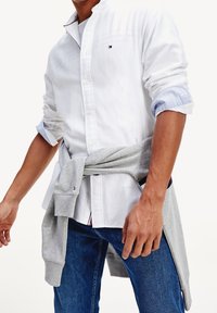 Camisa blanca de botonadura con puños a rayas azules, sudadera gris claro anudada a la cintura y jeans azul oscuro. Look casual y en capas.