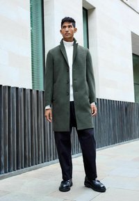 Grüner Wollmantel mit hohem Kragen und schwarzen Knöpfen, kombiniert mit einem weißen Rollkragenpullover und lockeren schwarzen Hosen; strukturierte Bodenfliesen.