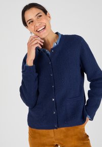 Cardigan navy in maglia con tessuto strutturato, colletto rotondo, chiusura con bottoni e due tasche frontali, indossato sopra una camicia di denim.