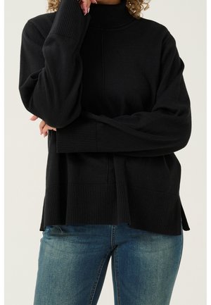 Persoon draagt een zwarte coltrui met geribbelde zoom en manchetten, gecombineerd met blauwe spijkerbroek, armen gekruist dichtbij het gezicht.