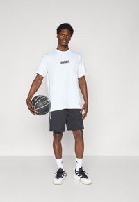 Bílá tričko s černým logem, černé basketbalové šortky, černé a bílé tenisky a bílé sportovní ponožky, držící černý basketbal.