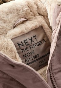 Brązowe sztuczne futro widoczne obok metki z napisem "NEXT THE FUTURE IS NOW EST.1982," z beżowym zewnętrznym materiałem i detalem zamka.