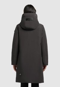 Juodas ilgas pūkinis paltas su dideliu gobtuvu, pagamintas iš švelnios medžiagos ir turintis stačiakampį logotipo ženklelį ant nugaros. Matomos šoninės kišenės.