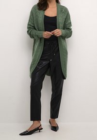 Jasnozielony sweter z dzianiny o fakturowanym wzorze, z otwartym przodem i długimi rękawami, w zestawie z czarnymi spodniami z sztucznej skóry i czarnymi czółenkami w szpic.