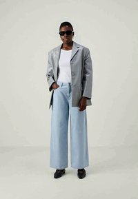 Blazer oversized cinza e brilhante por cima de uma camisa branca, combinado com jeans de perna larga azul-claro e sapatos pretos; modelo usa óculos de sol pretos.