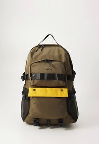 Olivzaļš mugursoma ar dzeltenu priekšējā kabatu, melnām siksnām un acu sānu kabatām; ir rāvējslēdzēji un logotips priekšpusē.