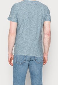 Pruhované modré tričko s krátkým rukávem, vyrobené z měkké tkaniny. Má kulatý výstřih a malé logo na levém rukávu. Kombinováno se světle modrými džíny.