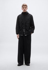 Must tuulekindel jope kõrge kraega, lukuga ja elastsete varrukaotstega. All näha ruuduline särk. Lahtised mustad püksid ja jalanõud.