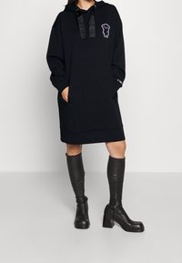 Schwarzes Hoodie-Kleid mit einer Vordertasche, oversized Silhouette und grafischem Akzent. Kombiniert mit knielangen schwarzen Blockabsatzstiefeln.