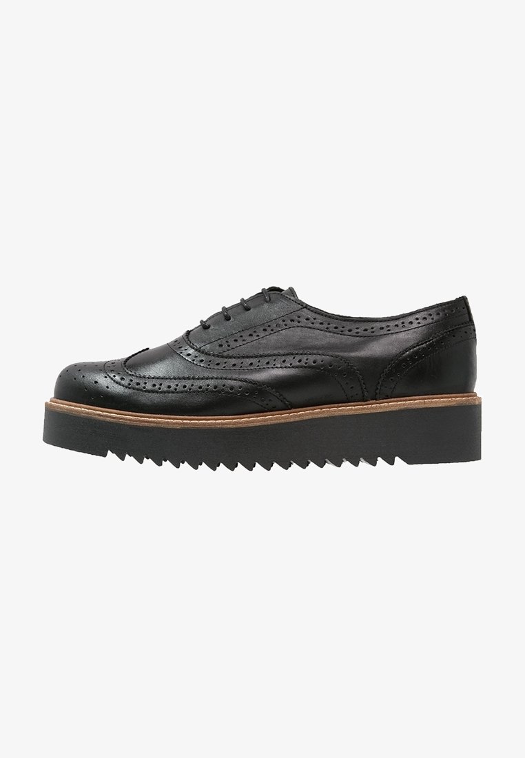 Chaussure brogue en cuir noir avec perforations en forme d'aile, semelle épaisse à bords festonnés et surpiqûres marron sur la trépointe, vue de côté sur fond blanc.