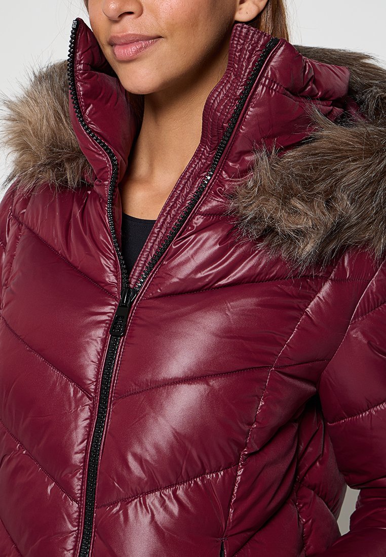 Vrouw in een glanzende kastanjebruine pufferjack met een hoge kraag en een met bont gevoerde capuchon, gedeeltelijk dichtgeritst over een zwarte top.