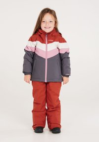Veste d'hiver colorée avec un motif en chevrons rose, blanc et gris, associée à un pantalon de ski rouge. Le matériau semble isolé et imperméable.