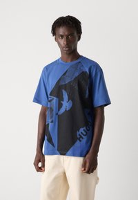 Zila kokvilnas T-kreklu ar melnu grafisko dizainu, kas ietver putnu un ģeometriskus formas, ar "HUGO" uzrakstu priekšpusē. Īsās piedurknes.