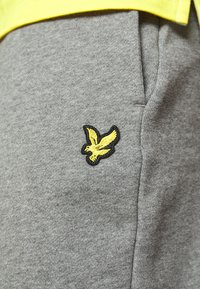 Grå joggebukser laget av myk bomullsmiks, med broderi av en gul fugl på låret og en glatt, teksturert stoffoverflate.