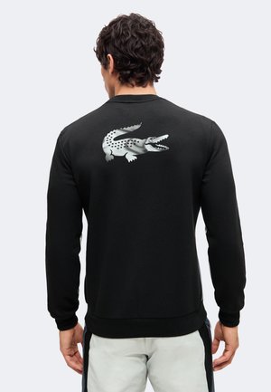 Homme de dos portant un sweat noir avec un grand logo crocodile argenté sur le haut du dos et un pantalon clair.