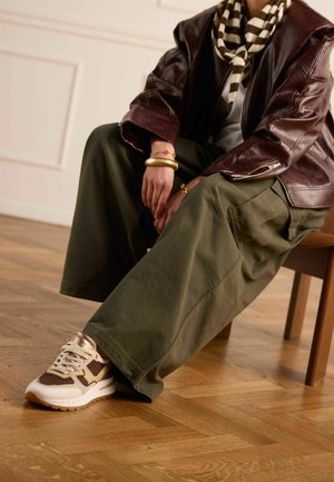 Personne assise sur une chaise en bois portant un pantalon large olive, des baskets marron et blanches, une veste en cuir marron, une écharpe rayée et des bracelets dorés.