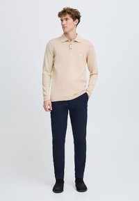 Beige hosszú ujjú póló, bordázott gallérral és mandzsettával, a mellkason egy kis logóval. Sötétkék, slim-fit nadrággal és sötétcipővel viselve.