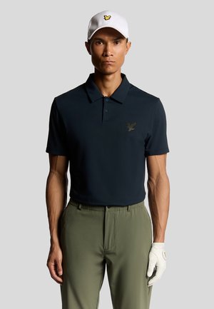 Marinblå polo t-shirt med korta ärmar och krage, med en diskret örnlogotyp. Matchad med olivgröna byxor och en vit keps.