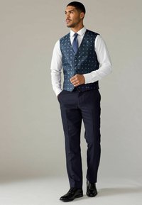Marineblaue Pünktchen-Weste über einem weißen Hemd, kombiniert mit dunklen Hosen und schwarzen Schnürschuhen. Die Weste hat ein maßgeschneidertes Design mit einer glatten Textur.