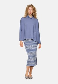 Helles blauer, langärmeliger Oberteil mit Rollkragen; gemusterter Midirock in Blau und Weiß mit kreisförmigen Motiven; graue, spitz zulaufende Schuhe.