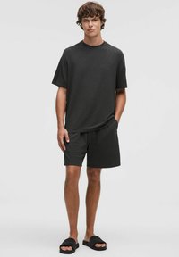T-shirt grigio scuro a maniche corte e pantaloni shorts abbinati realizzati in tessuto morbido. Il modello indossa sandali neri con cinturino. Design semplice e dalla vestibilità rilassata.