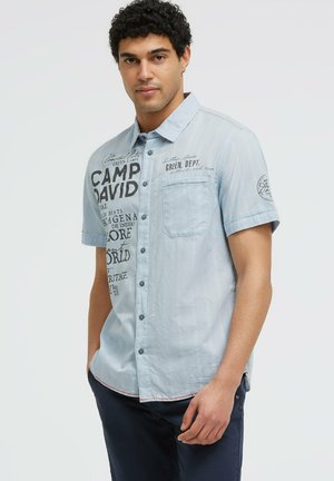 Mann mit lockigem Haar, der ein hellblaues kurzärmeliges Hemd mit schwarzem Textdruck und dunkelblaue Hose trägt.