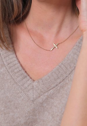 Femme portant un pull en maille beige à col en V, montrant un pendentif croix en or sur une chaîne délicate autour du cou.