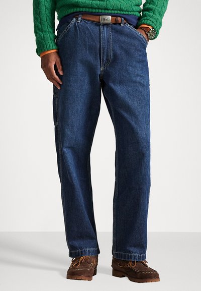 Dunkelblaue Jeans aus Denim mit lockerer Passform, fünf Taschen und kontrastierenden Nähten. Kombiniert mit braunen Schuhen und einem grünen Strickpullover.