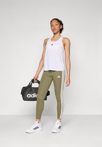 Canottiera sportiva bianca con logo nero, leggings verde oliva con strisce bianche e borsa a tracolla nera con logo bianco.