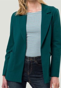 Femme portant un blazer vert foncé sur une chemise blanche à rayures bleu sarcelle, jean bleu foncé, cadrée aux épaules et à la taille.