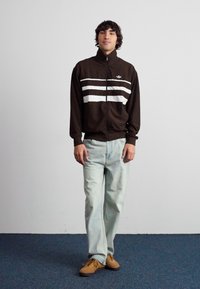 Καφέ τζάκετ με φερμουάρ και λευκές ρίγες, με υφασμάτινο σχέδιο. Συνδυασμένο με ανοιχτό μπλε τζιν και καφέ παπούτσια σε μπλε χαλί.