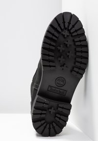 Musta kumisaappaan pohja syvällä uramallella, jossa on pyöreä logo ja teksturoituja yksityiskohtia. Kestävä muotoilu vahvistetuilla reunoilla.