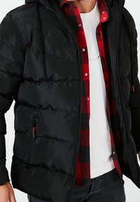 Chaqueta negra acolchada con capucha, con diseño acolchado y bolsillo con cremallera. Combinada con una camisa de franela a cuadros roja y negra debajo.