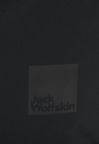 Schwarze Stofftasche mit einem rechteckigen Patch, auf dem das geprägte Logo "Jack Wolfskin" in Dunkelgrau zu sehen ist. Die Textur ist gewebt und robust.