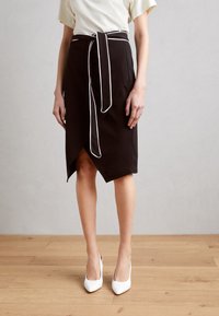 Černá zavinovací sukně s bílým lemováním, délka ke kolenům, s zavázáním v pase. V kombinaci s bílými lodičkami na dřevěné podlaze.