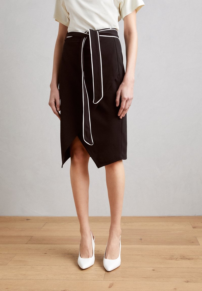 Černá zavinovací sukně s bílým lemováním, délka ke kolenům, s zavázáním v pase. V kombinaci s bílými lodičkami na dřevěné podlaze.