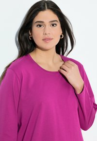 Fuchsia lange mouwen shirt met een ronde hals, gemaakt van een gladde stof. Model heeft lang donker haar en houdt met één hand de halslijn vast.